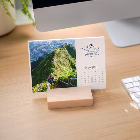 Desk calendar card displayed on a desk, featuring a person hiking steep mountain stairs with lush green ridges and a distant city below.