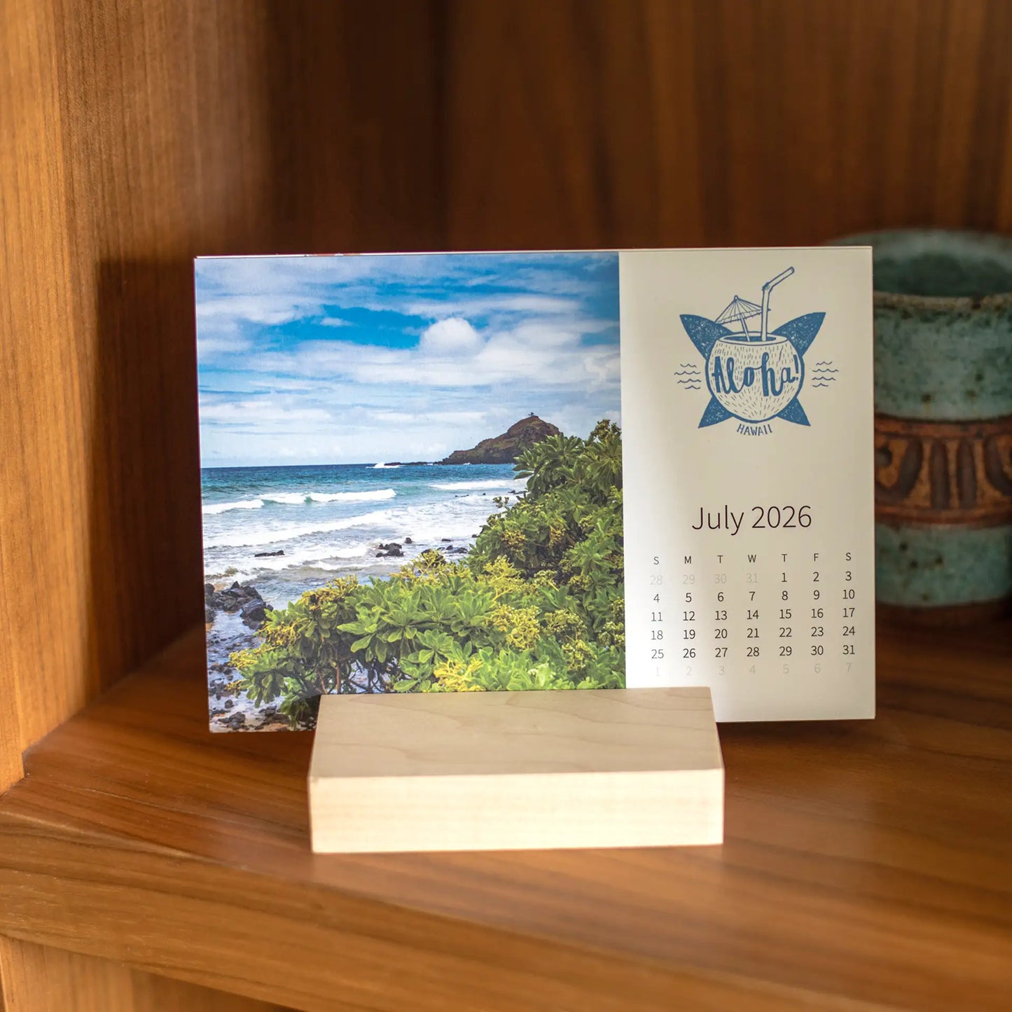 Desk calendar card in a wooden stand showing a tropical coastline with ocean waves, rocky shore, green foliage, and a bright blue sky.