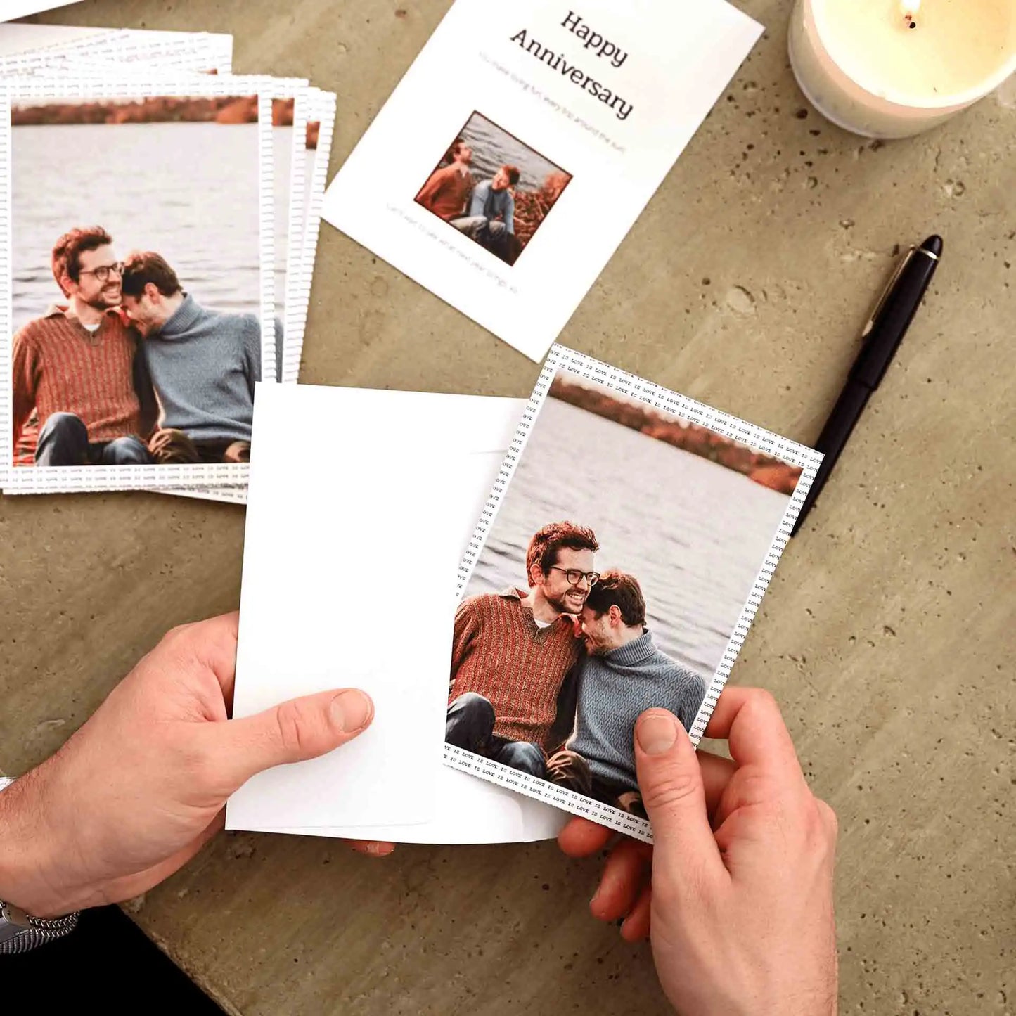 Flat card with “Happy Anniversary” text and small photo, surrounded by printed photo cards, envelope, pen, and candle on table.