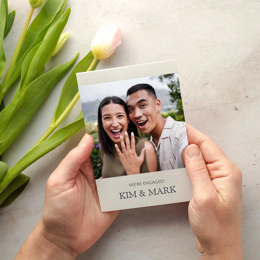 Flat card held in hands featuring smiling couple photo with engagement announcement text and names printed below, next to tulip flowers.