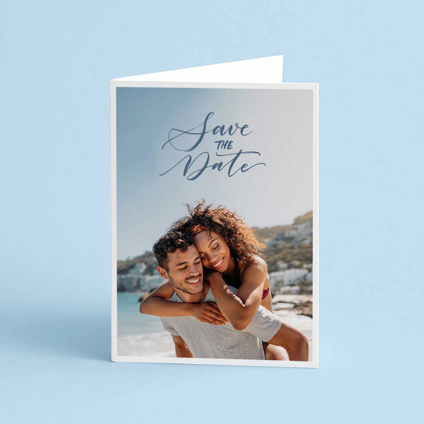 Folded greeting card with “Save the Date” text and photo of a couple embracing on a beach, set against a light blue background.