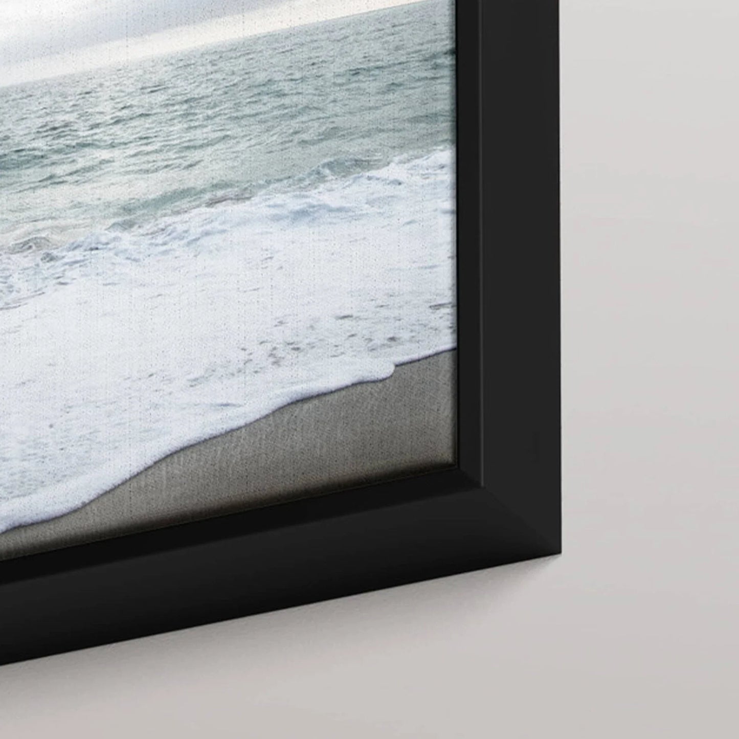 Black framed canvas print featuring ocean waves rolling onto sandy shore, shown in close-up detail against a light wall.