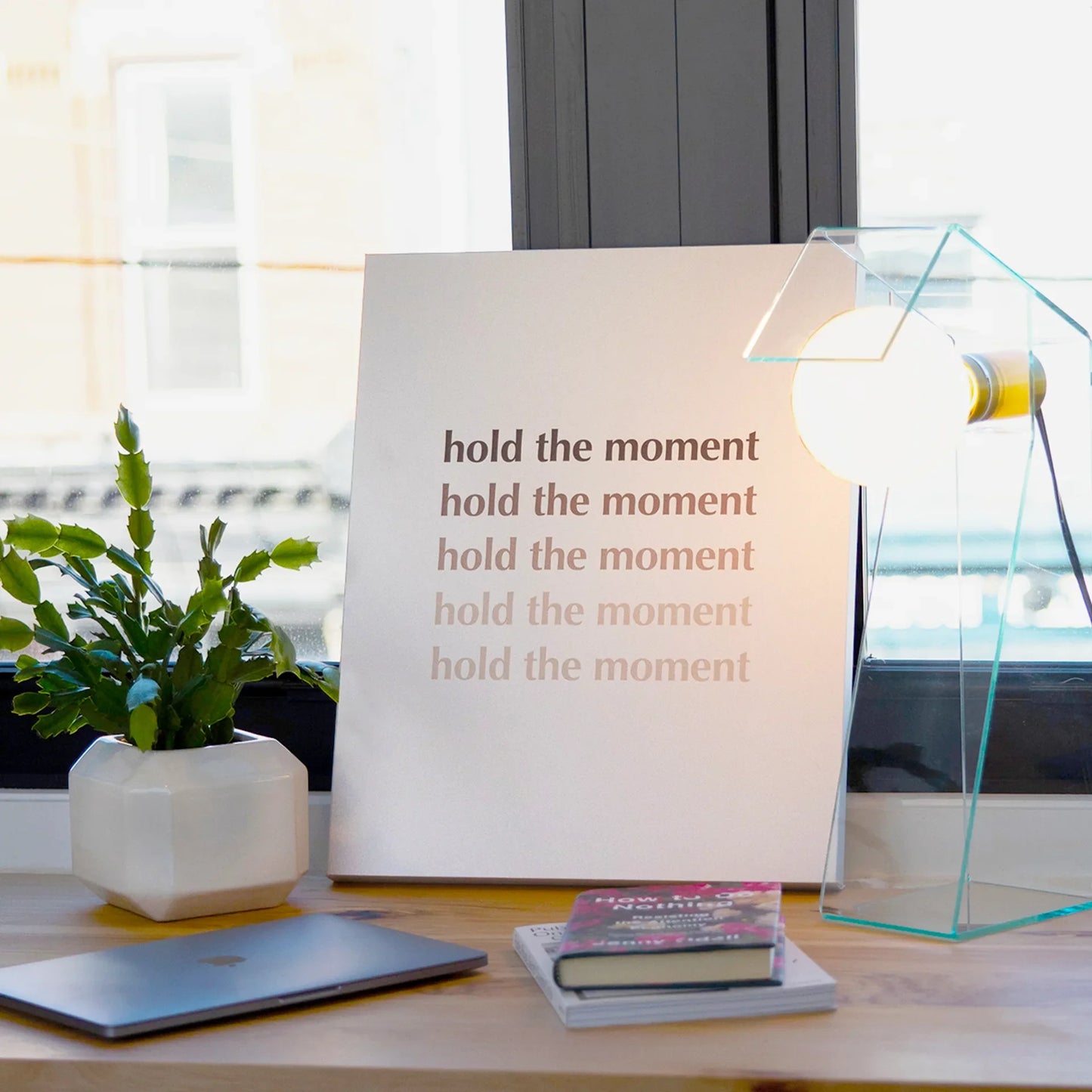 Gallery wrapped canvas featuring repeated “hold the moment” typography in neutral tones, displayed on a desk beside a glass lamp and potted plant.