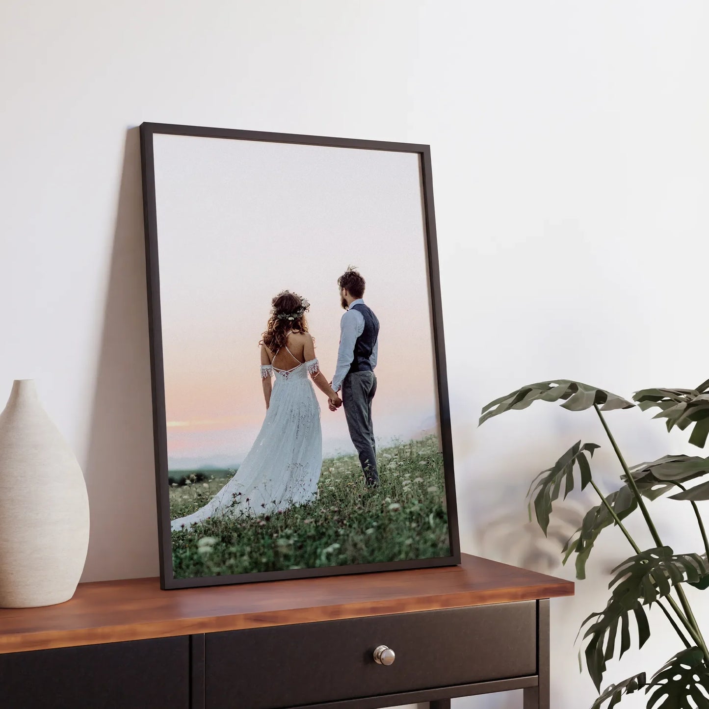Black framed large-format vertical photo print of a couple holding hands in a grassy field at sunset, displayed on a wooden console beside a vase and plant.