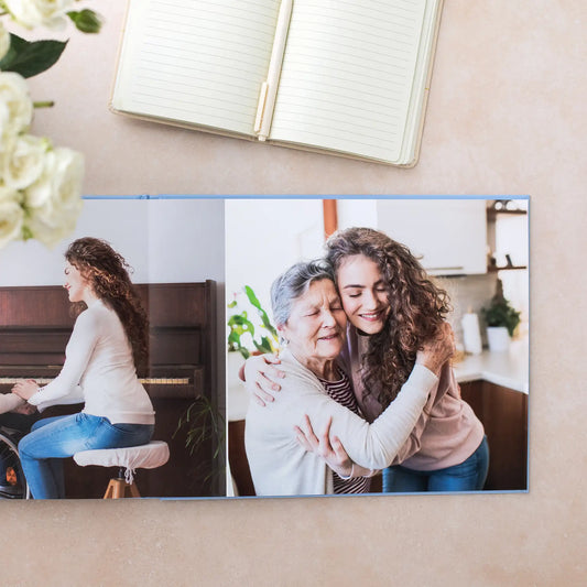Layflat photo book open to two-page spread with portraits of a younger person and an older person embracing, placed beside notebook and flowers.