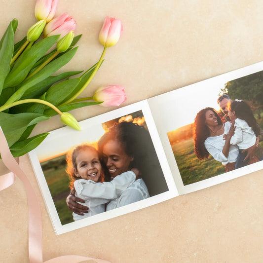Open softcover photo book showing a parent and child embracing outdoors at sunset, styled with tulips and ribbon on a light surface