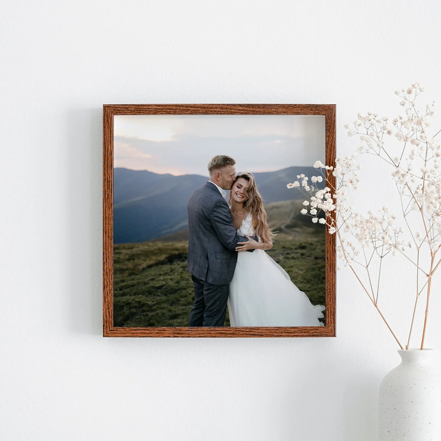 Framed square photo print showing a couple embracing in a mountain landscape, displayed on a wall next to a decorative vase.