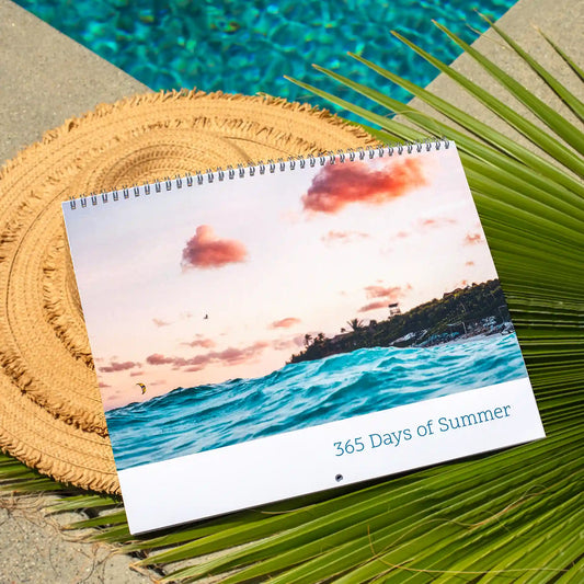 Wall calendar cover showing ocean waves at sunset with pink clouds, text reads “365 Days of Summer,” placed on woven mat with palm leaves.