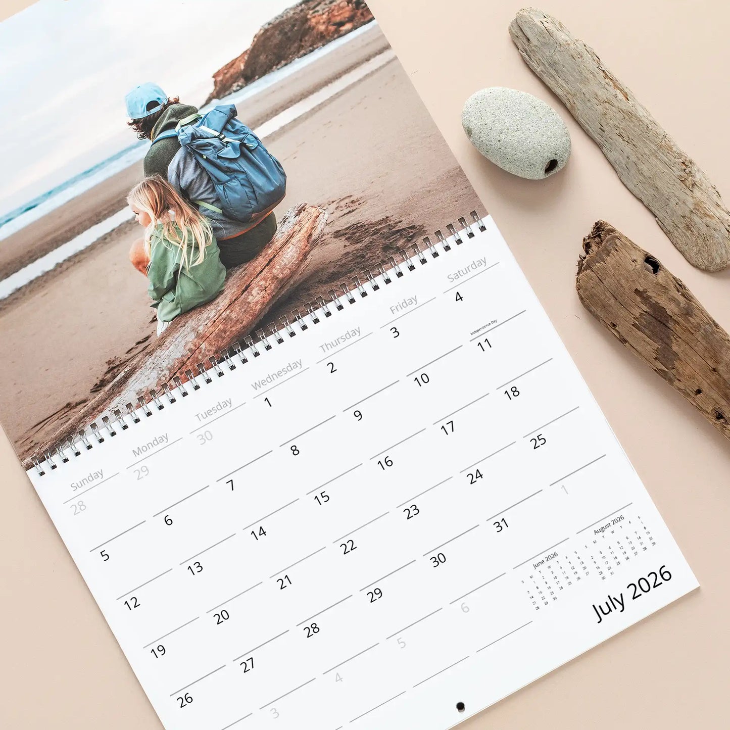 A wall calendar laid flat showing an adult and child sitting on driftwood at a beach, with spiral binding and July 2026 grid visible.