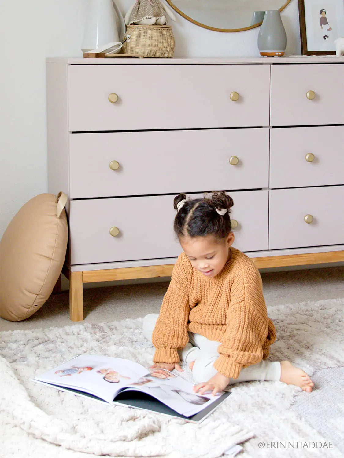Young child sitting on a soft rug in a nursery reading a large photo book, with a dresser, woven basket, and neutral decor behind.
