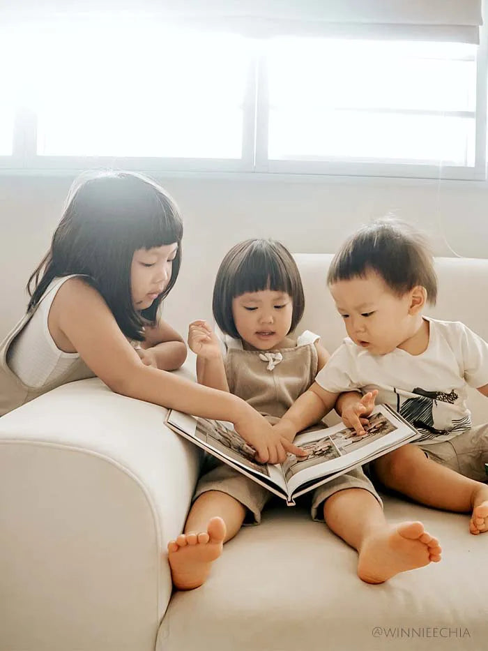 Three young children sitting together on a light-colored couch looking through a hardcover photo book with printed family pictures in a bright living room.