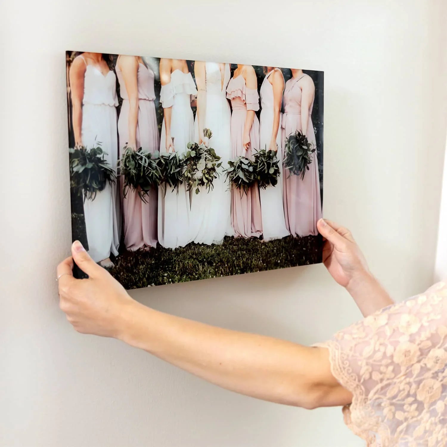 Acrylic wall print of bridesmaids in blush dresses holding green bouquets, being positioned on a light-colored wall.