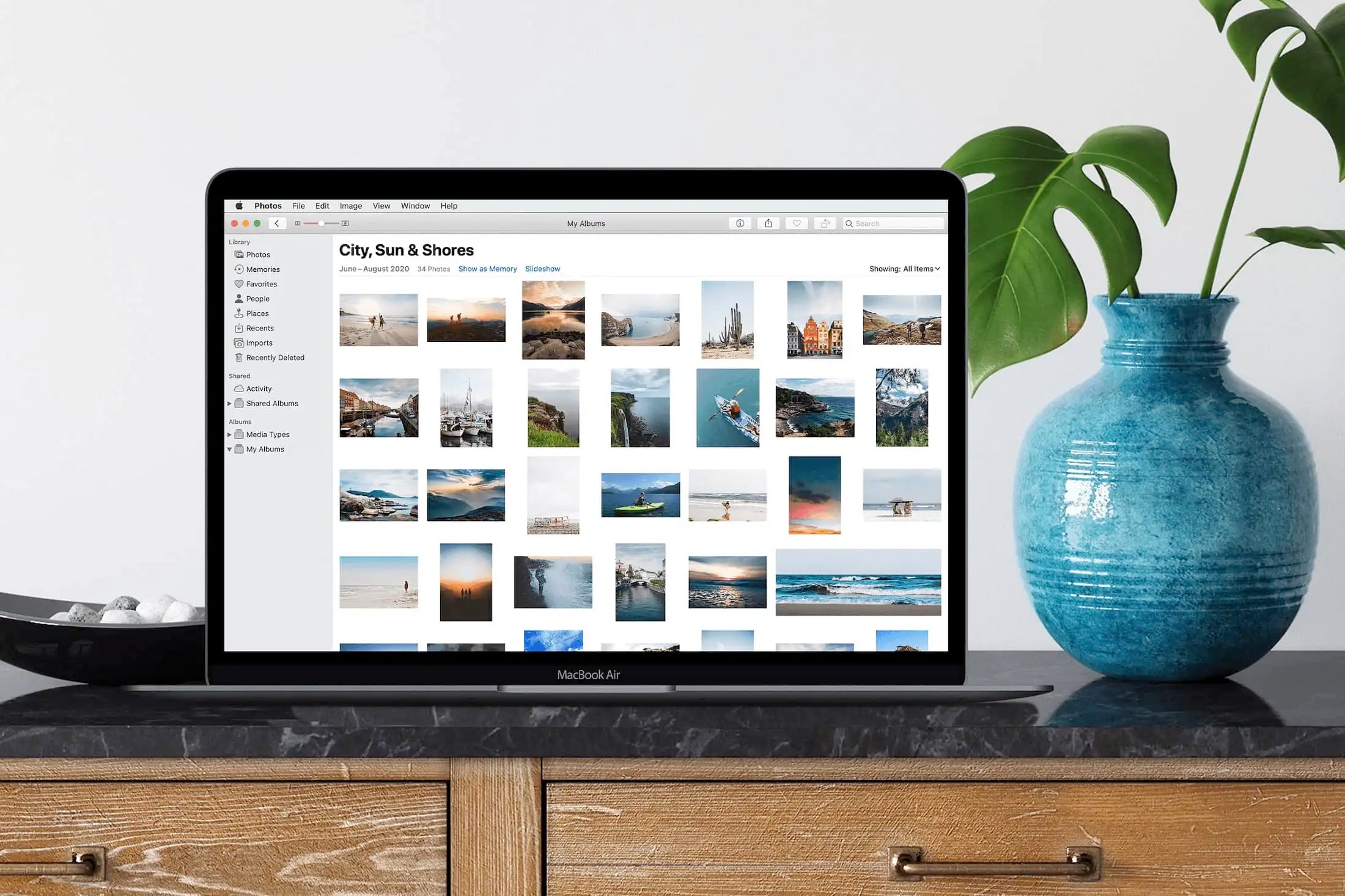 MacBook Air displaying Apple Photos travel album with grid of beach and landscape images, placed on console table beside blue ceramic vase.