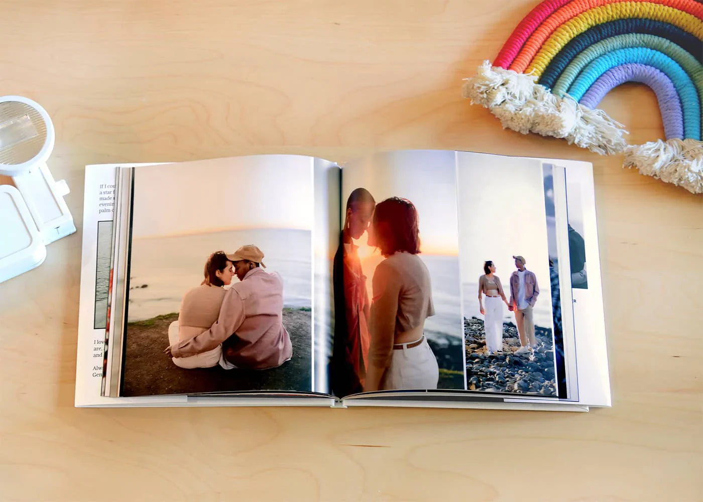 Open hardcover photo book displaying romantic couple photographs across multiple pages, resting on wooden table with rainbow decoration nearby