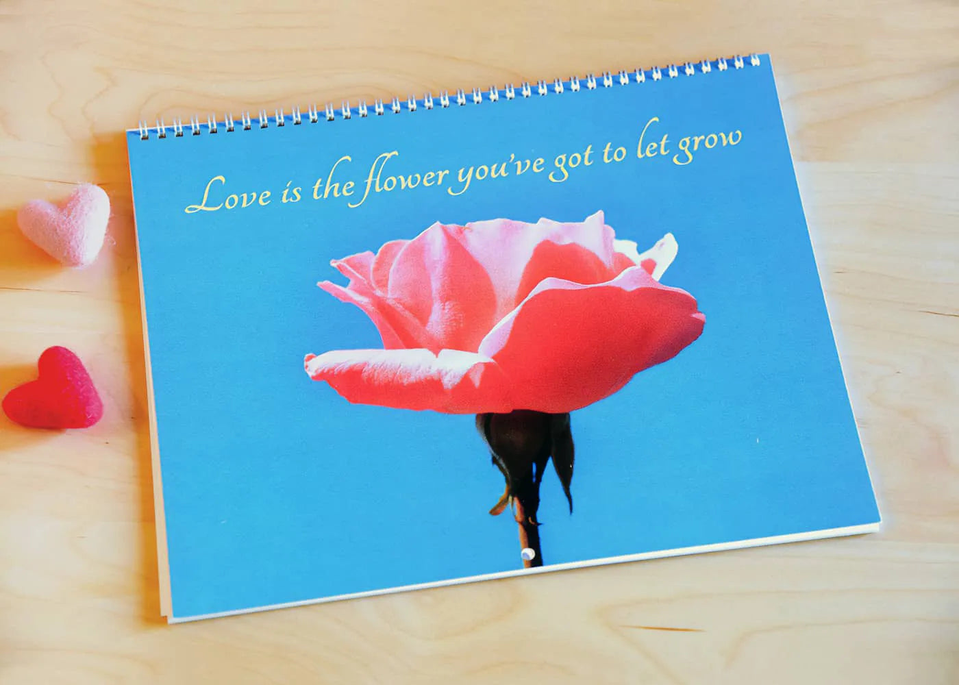 Spiral-bound photo calendar with red rose cover and printed quote displayed on wooden table with small decorative heart accents.