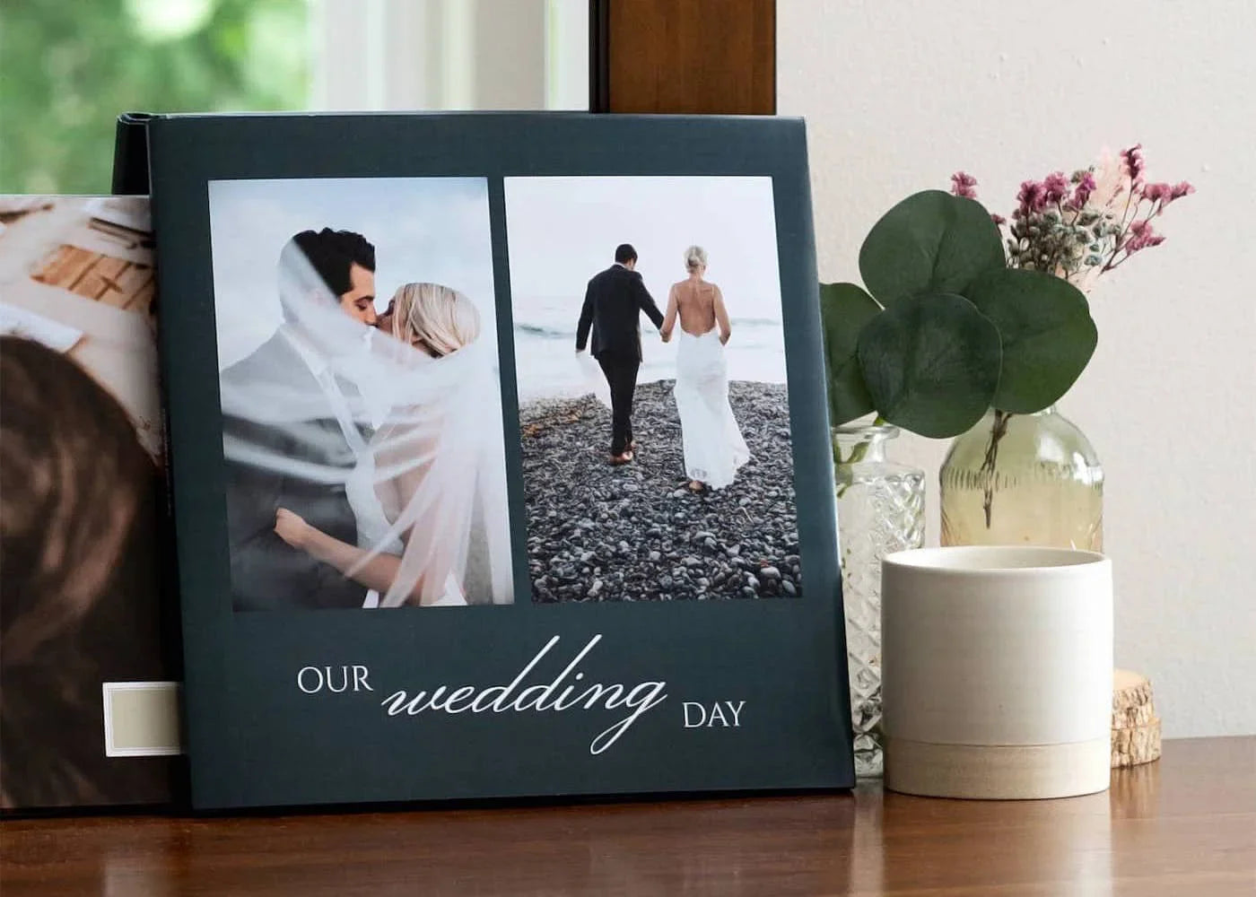 Hardcover wedding photo book with couple portraits and title “Our Wedding Day” displayed on table beside candles and small glass vase.