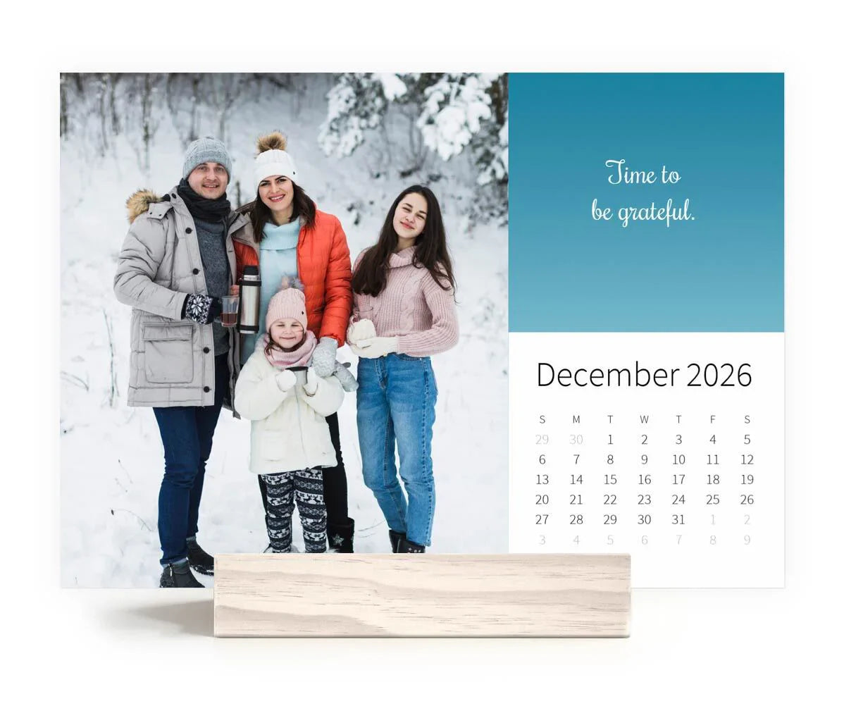 Desk calendar on wooden stand featuring a winter family portrait in the snow with the quote “Time to be grateful” and December 2026 monthly grid.