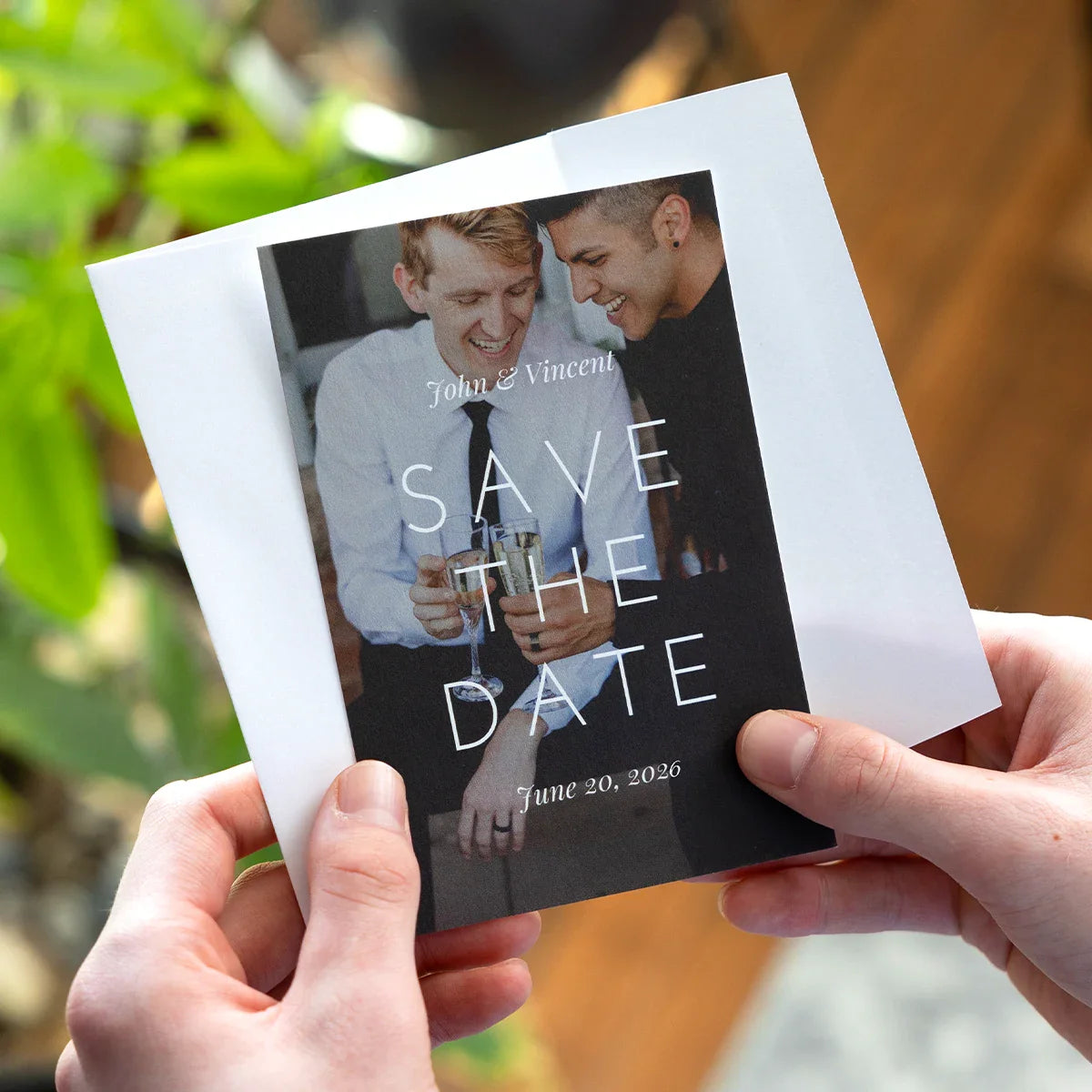 Flat save-the-date card held in hands featuring a photo of two men smiling and holding champagne glasses, with text reading “John & Vincent – Save the Date – June 20, 2026.”