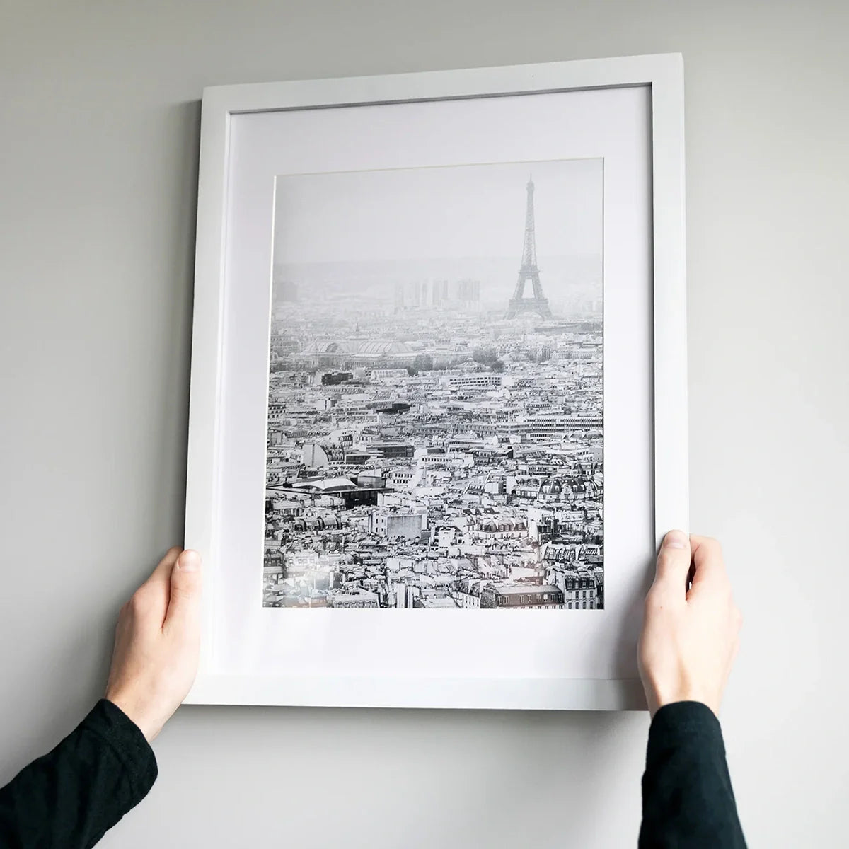 White-framed matte print of a black-and-white Paris cityscape with the Eiffel Tower, being hung on a wall.