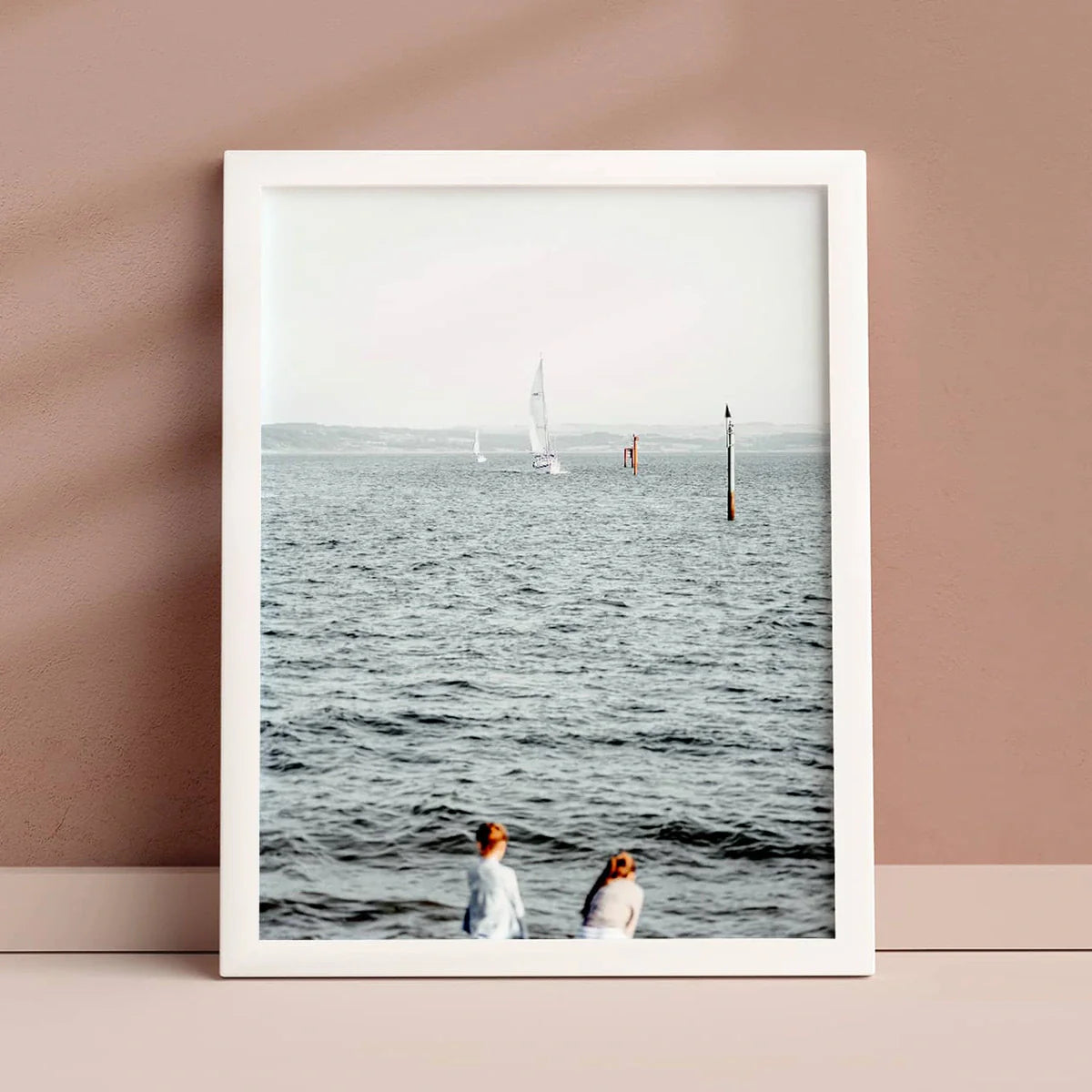 White-framed print of sailboats on the water with two people in the foreground, displayed against a soft pink wall.