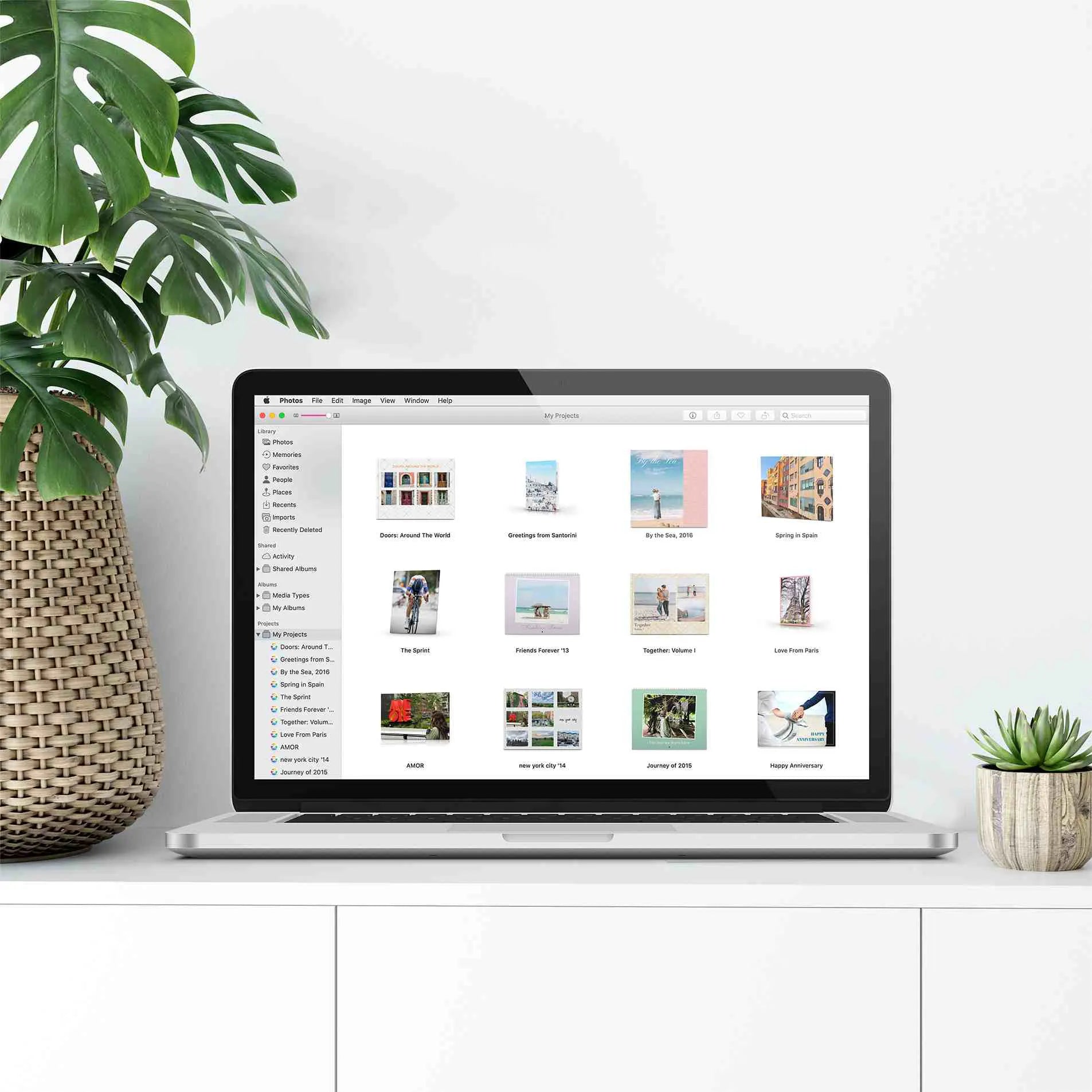 Laptop on a white cabinet displaying Apple Photos projects library with multiple photo book and print project thumbnails, surrounded by houseplants in a minimalist home setting.