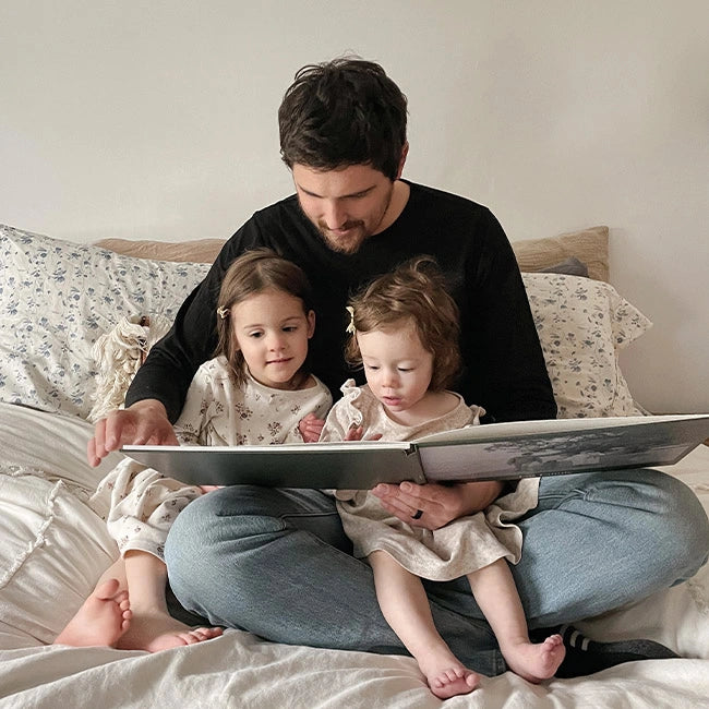 Man showing a photo book to two young children on a bed.