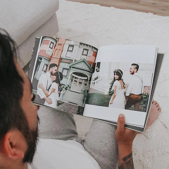 A man sitting on the floor flipping through a hardcover photo book filled with couples portrait photos.