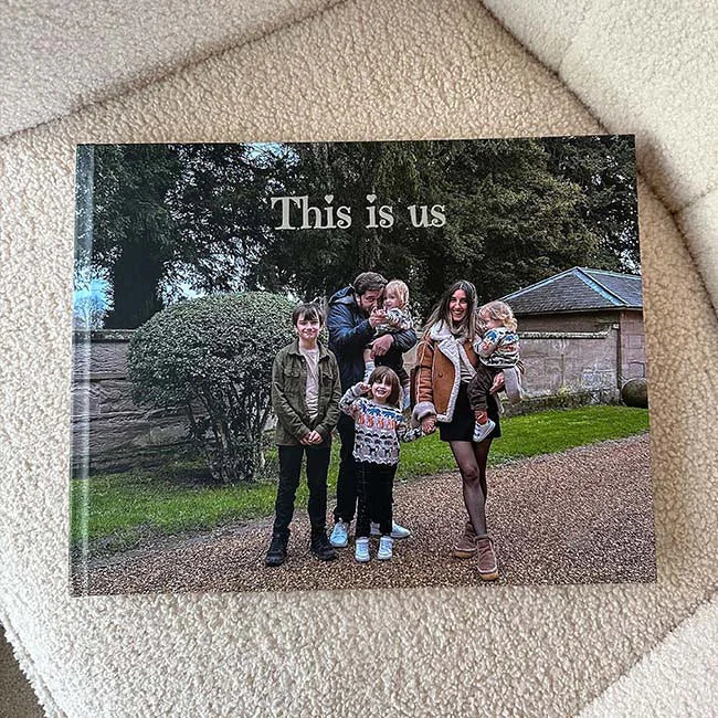 A custom hardcover photo book resting on a cozy cream sherpa surface, featuring a family portrait on the cover with the personalized title "This is us." The cover photo shows a family of five — two parents and three young children — standing together outdoors on a gravel path surrounded by lush greenery and stone walls.