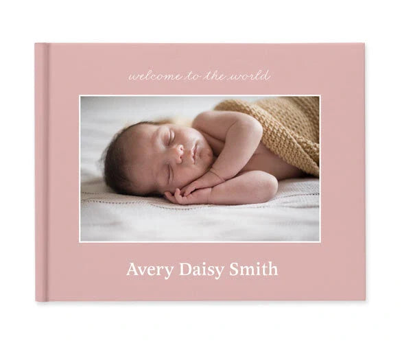 Layflat baby photo book cover in blush pink with centered newborn portrait, scripted title text, and custom name printed below.