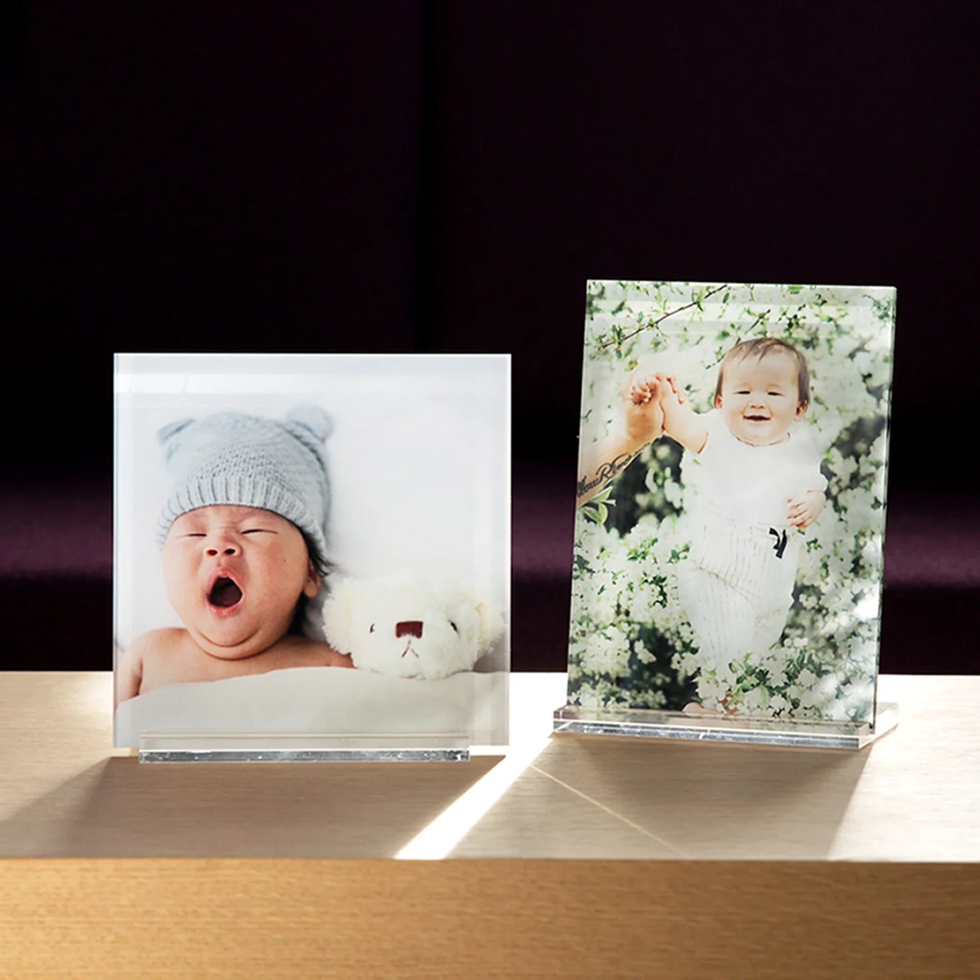 Two acrylic desk prints displayed side by side on a wooden surface, featuring a newborn portrait and a smiling baby in blooming flowers.