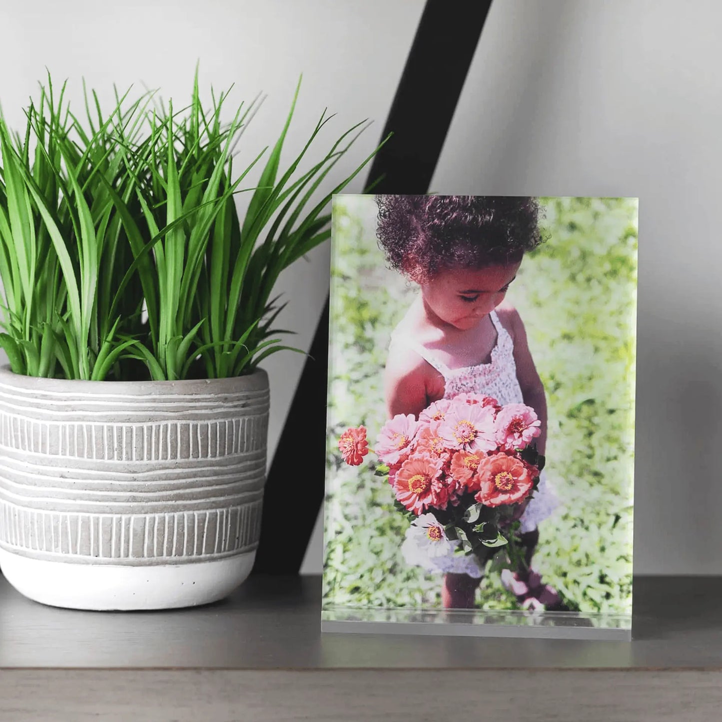 Acrylic desk print featuring a young child holding a bouquet of pink flowers, styled on a tabletop beside a textured plant pot.