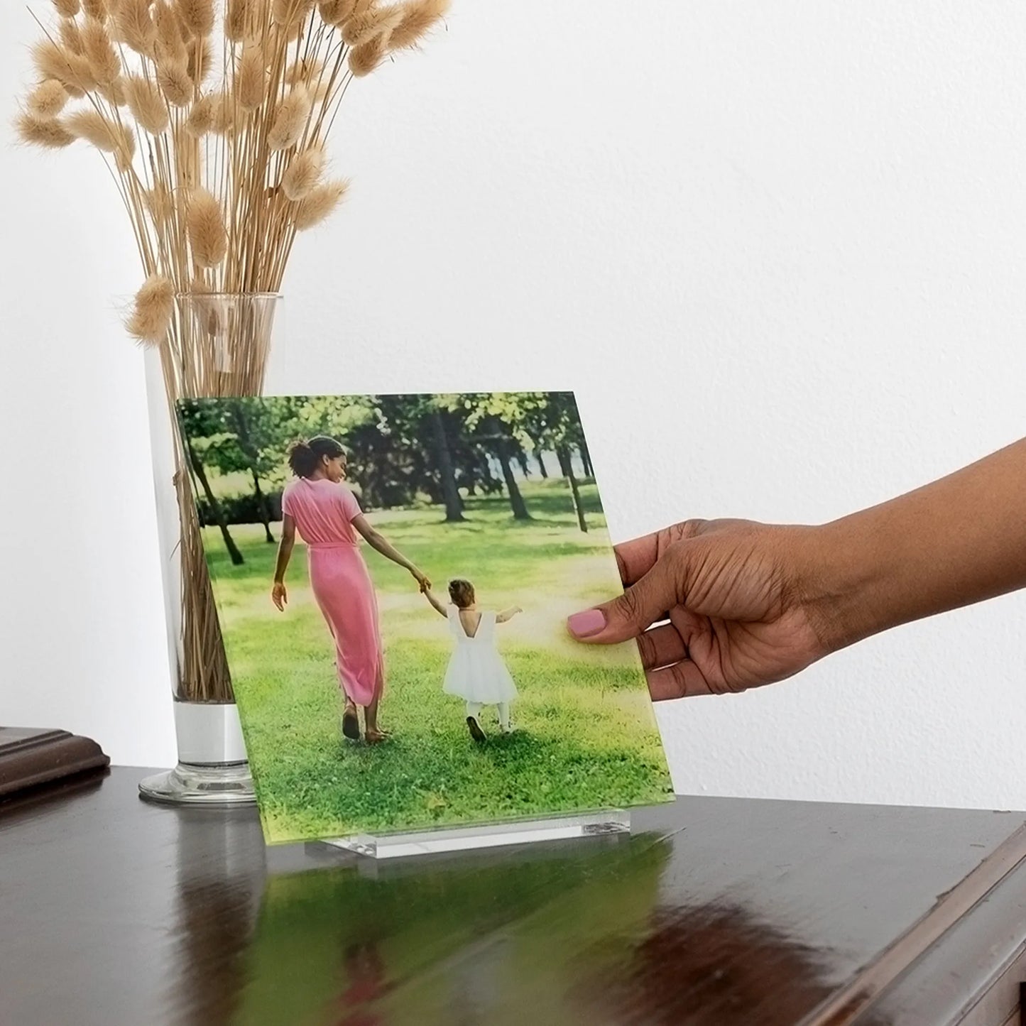 Acrylic desk print of a mother in a pink dress holding her child’s hand in a sunny park, shown on a dark wood table with dried florals.
