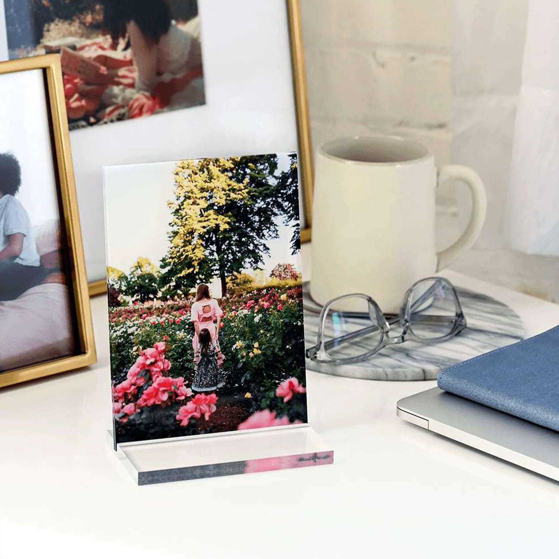 Acrylic desk print featuring a mother and daughter walking through a colorful flower garden, displayed on a white desk beside framed photos, glasses, and a laptop.