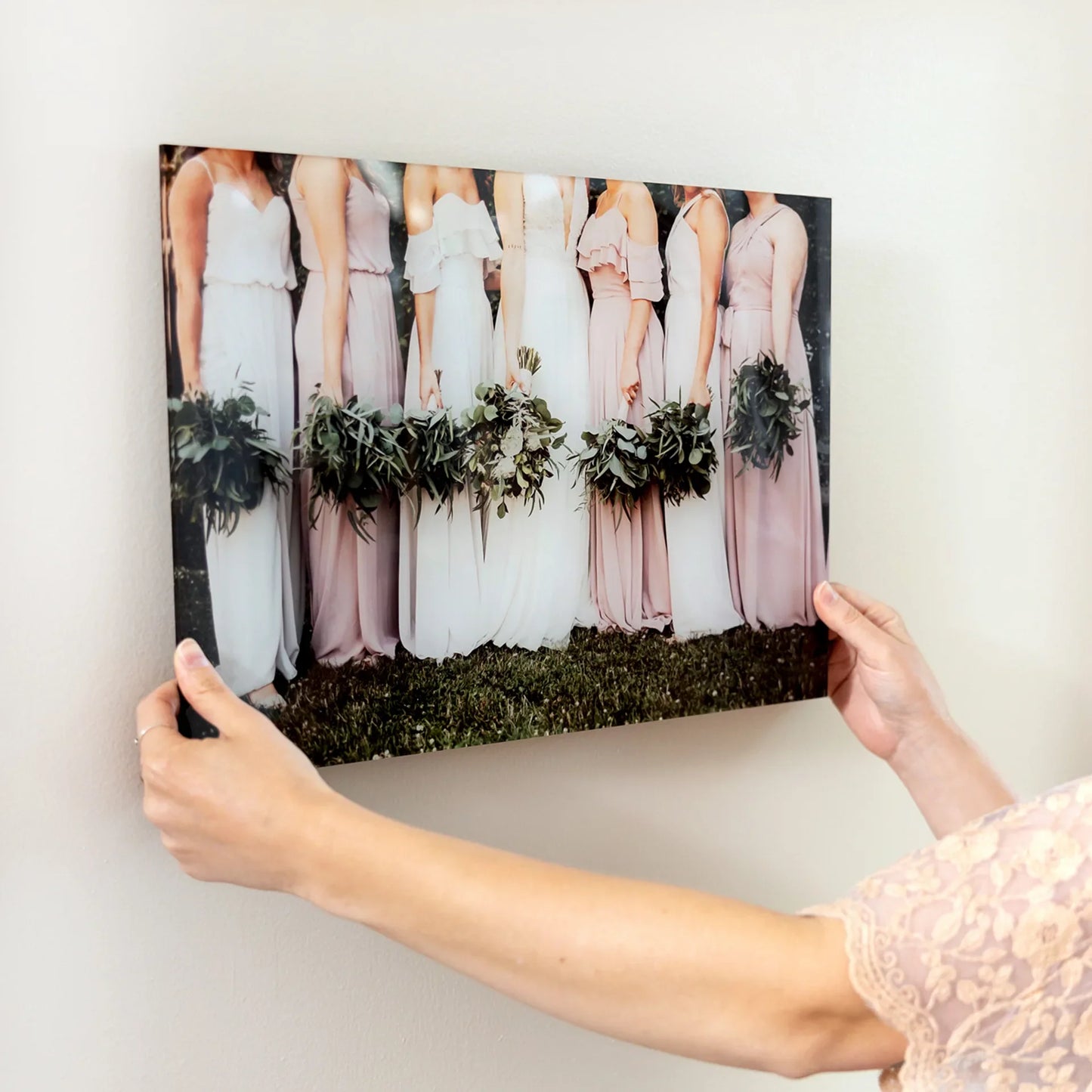 Acrylic wall decor print of bridesmaids in soft-toned dresses holding greenery bouquets, being positioned on a light-colored wall.