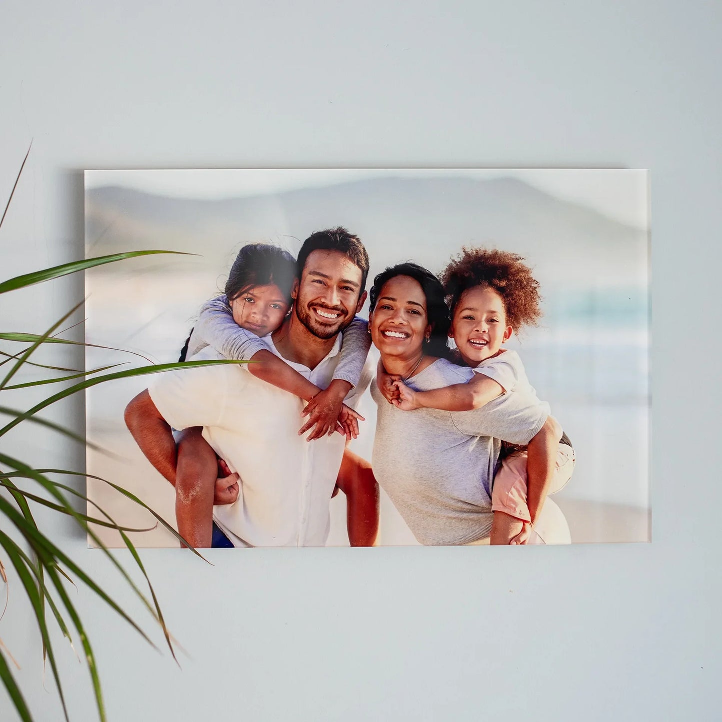 Acrylic wall decor print featuring a smiling family of four posing together outdoors with mountains in the background, mounted on a light-colored wall.