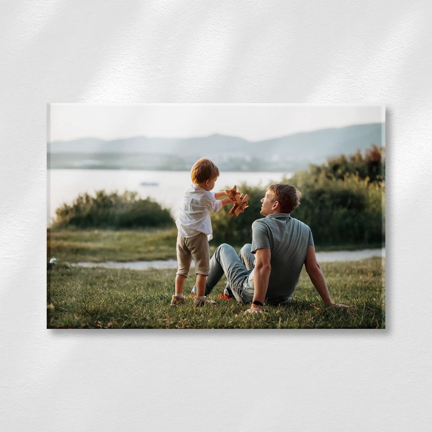 Acrylic wall print featuring a person sitting on grass with a child holding a toy airplane near a lake, with soft mountains in the background.