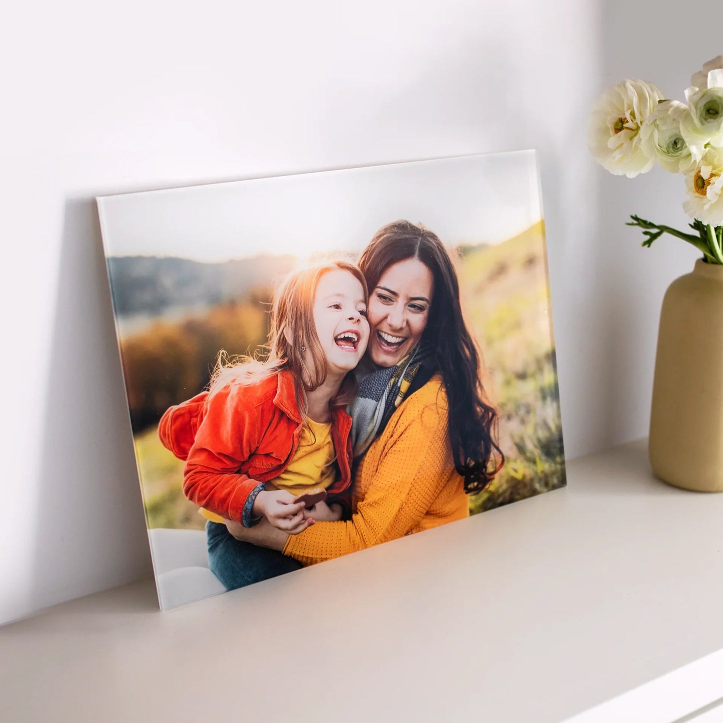 Acrylic wall decor print of a woman and young child laughing together outdoors in warm sunlight, displayed on a white shelf beside flowers.