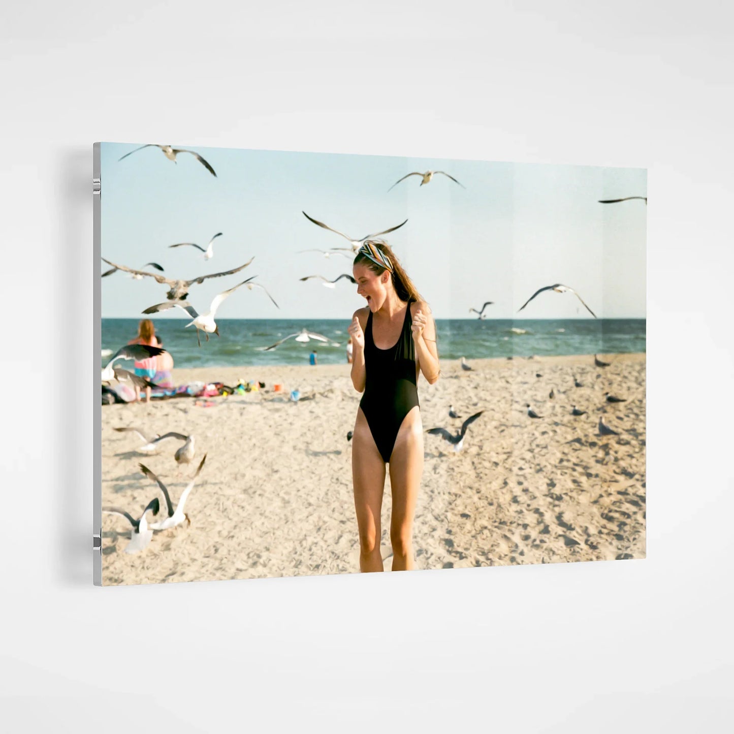 Acrylic wall decor print of a woman in a black swimsuit standing on a sandy beach surrounded by flying seagulls, displayed on a white wall.