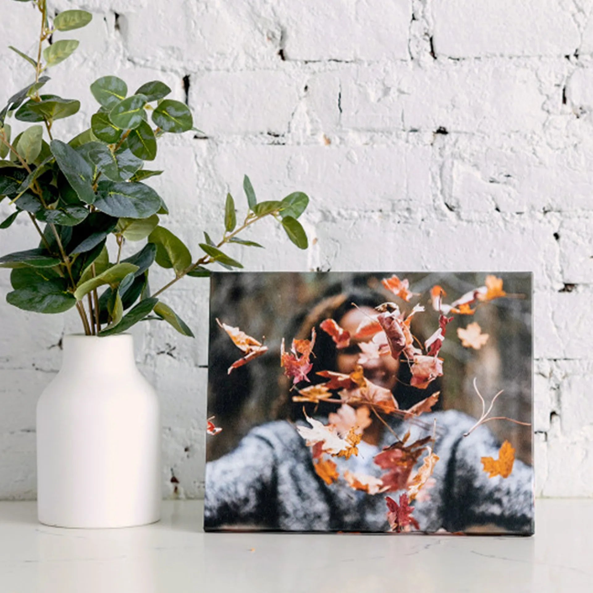 Canvas desk print of a person tossing orange and red autumn leaves, displayed on a white surface against a painted brick wall.