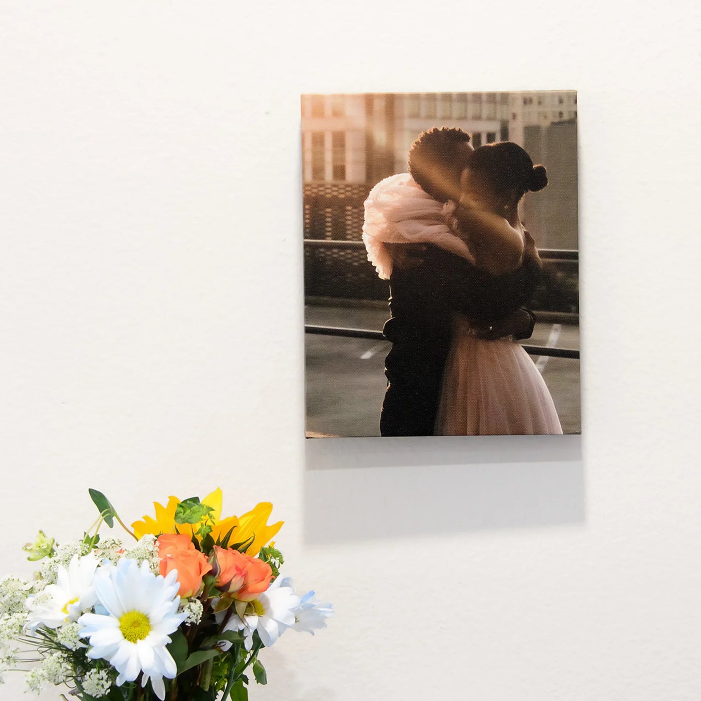 Canvas desk print featuring a couple embracing at sunset on a rooftop, displayed on a white wall above a vase of mixed flowers.