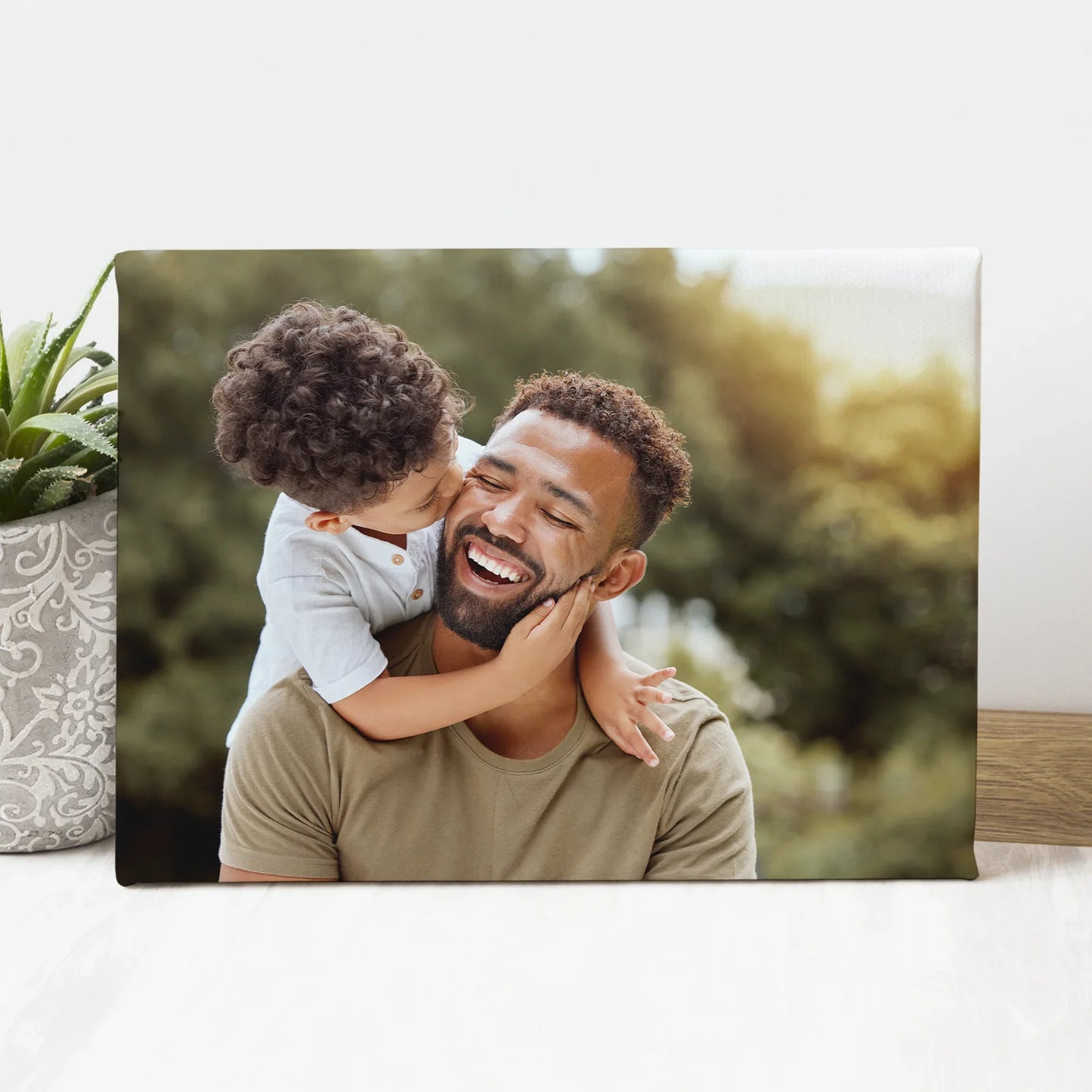 Canvas desk print featuring a child kissing a smiling father on the cheek outdoors, styled on a white tabletop beside a potted plant.