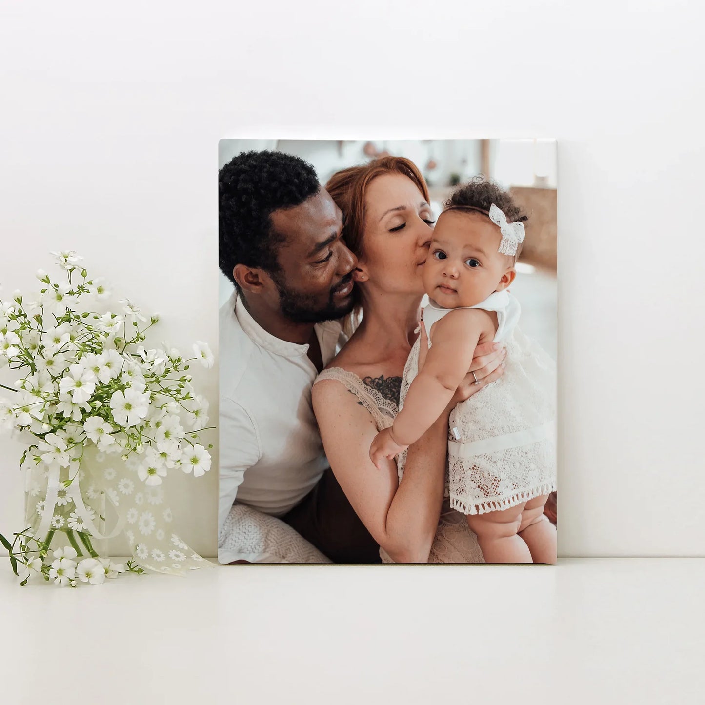 Canvas desk print of two parents holding and kissing a baby wearing a white headband, displayed beside a glass vase of white flowers.