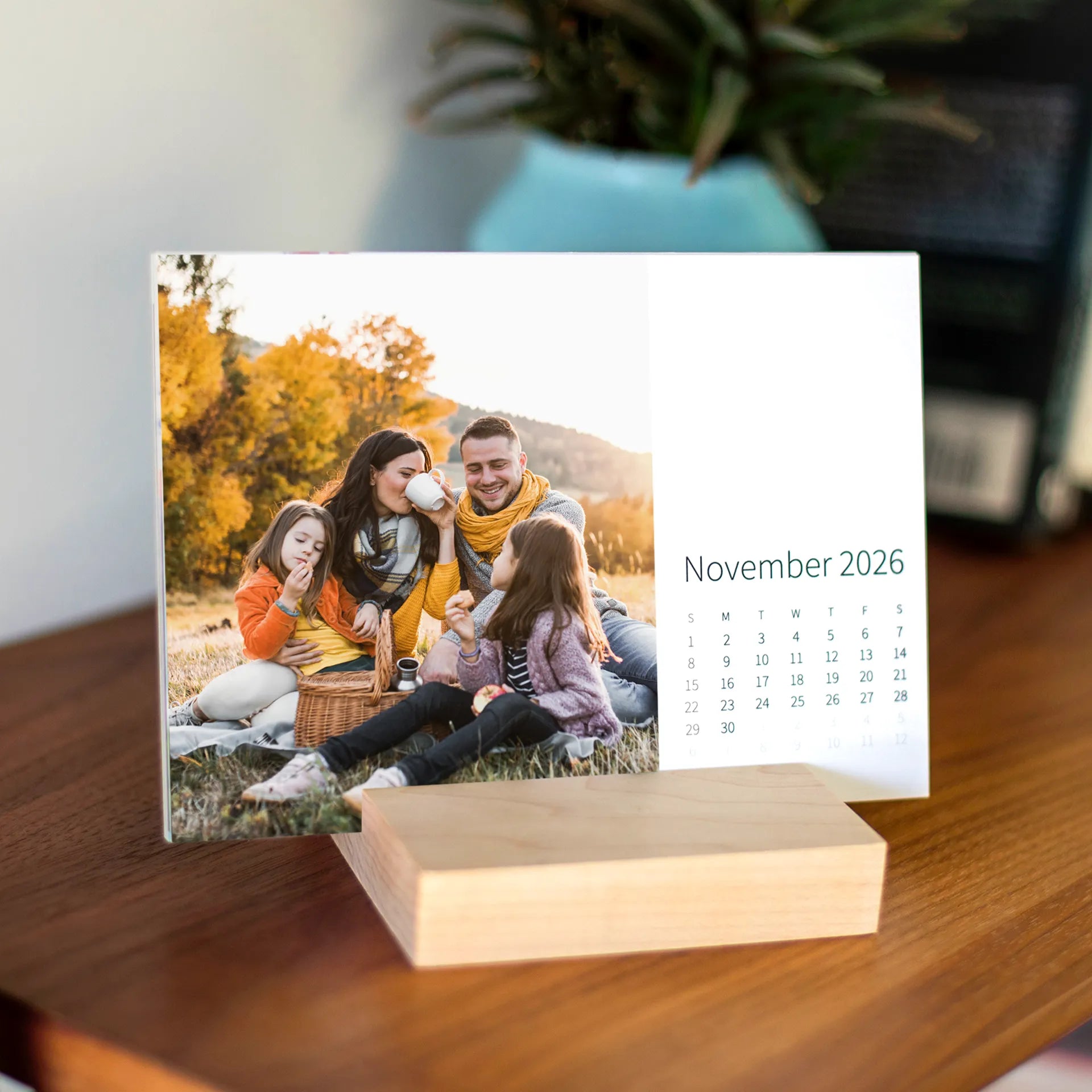 Desk calendar card displayed in a wooden base, featuring a family sitting together during an autumn picnic with trees and warm fall colors.