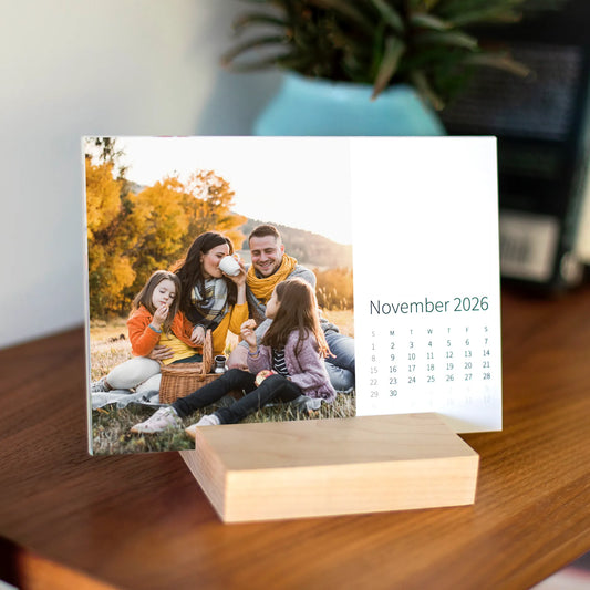 Desk calendar card displayed in a wooden base, featuring a family sitting together during an autumn picnic with trees and warm fall colors.