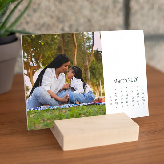 Desk calendar card displayed in a wooden stand, showing a parent and child sitting on a blanket in a park surrounded by trees.