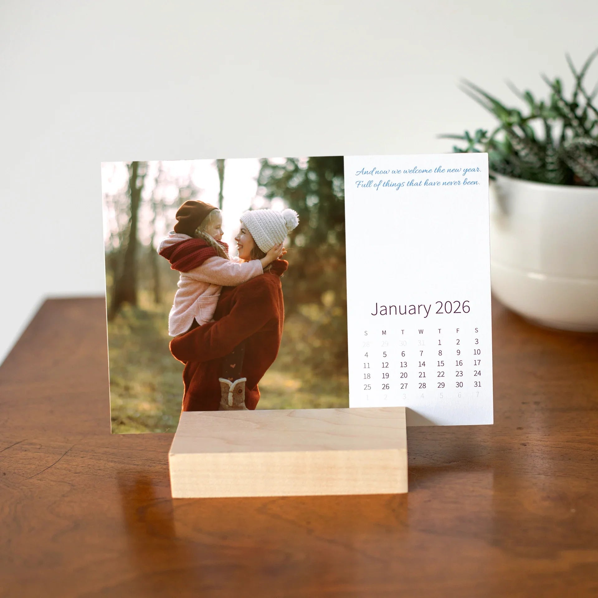 Desk calendar card displayed in a wooden base, showing a parent holding a child outdoors in winter clothing with soft natural light.