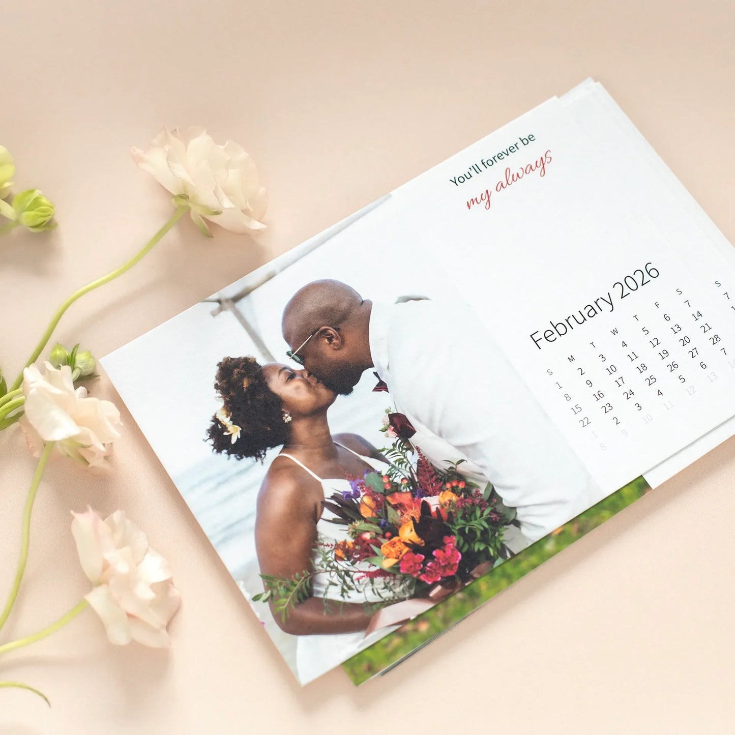 Desk calendar card lying flat, featuring a couple in wedding attire kissing outdoors, holding a bouquet, with flowers arranged nearby.