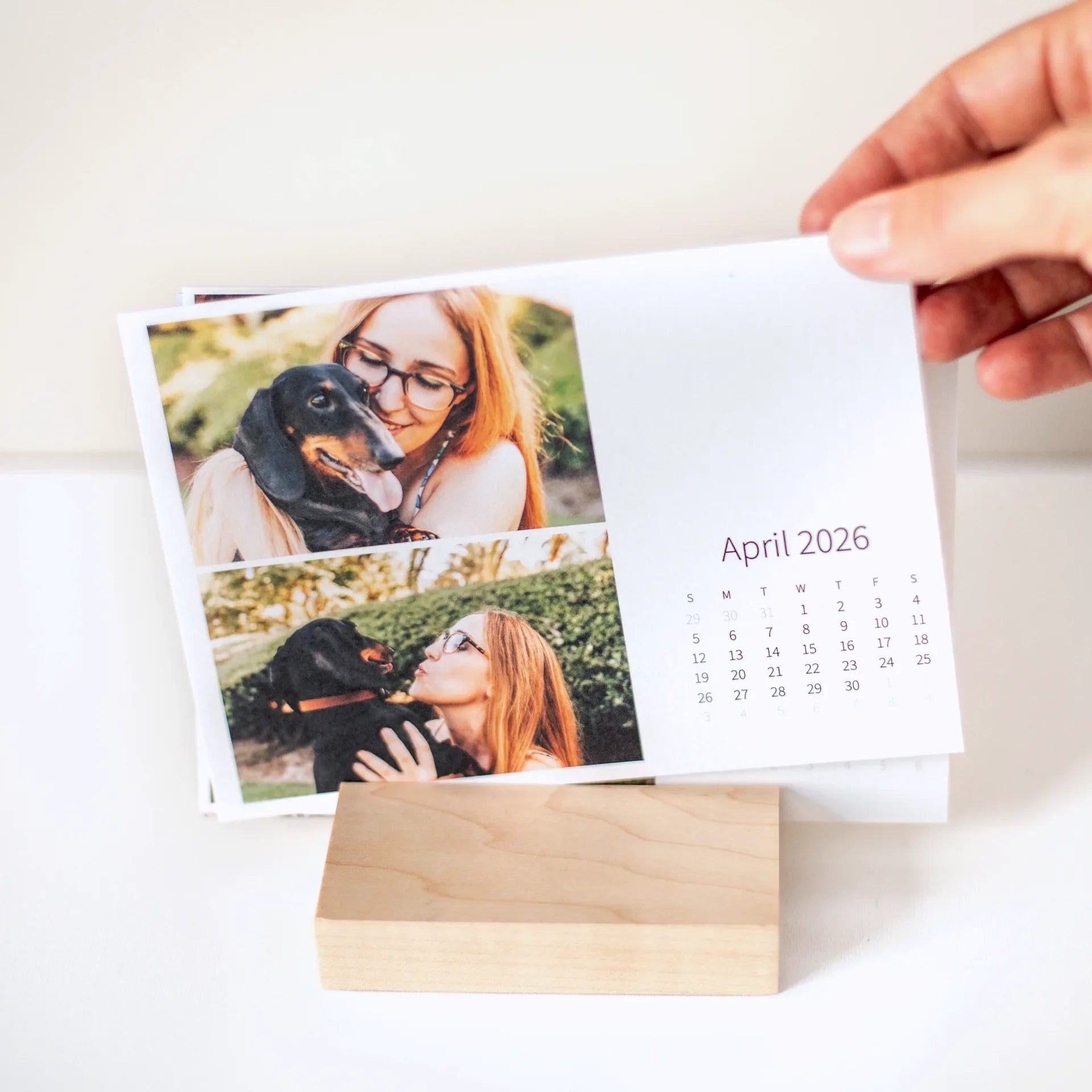 Desk calendar card held above a wooden base, featuring a woman with glasses cuddling a small black dog outdoors in natural light.