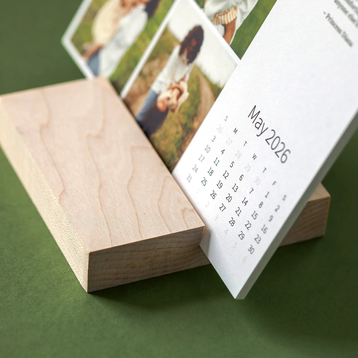 Close-up of a desk calendar card resting in a smooth wooden base, showing the month layout and layered photo cards behind.