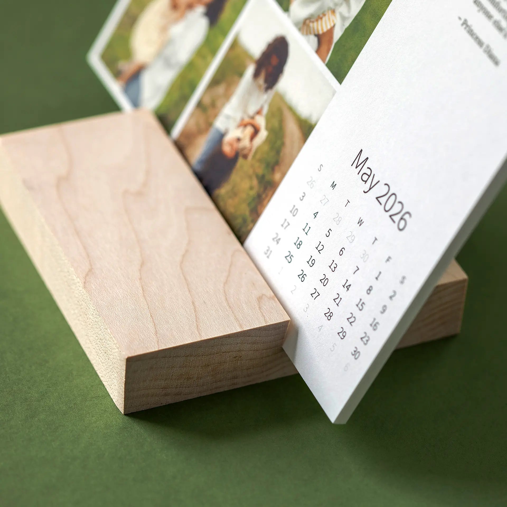 Close-up of a desk calendar card resting in a smooth wooden base, showing the month layout and layered photo cards behind.